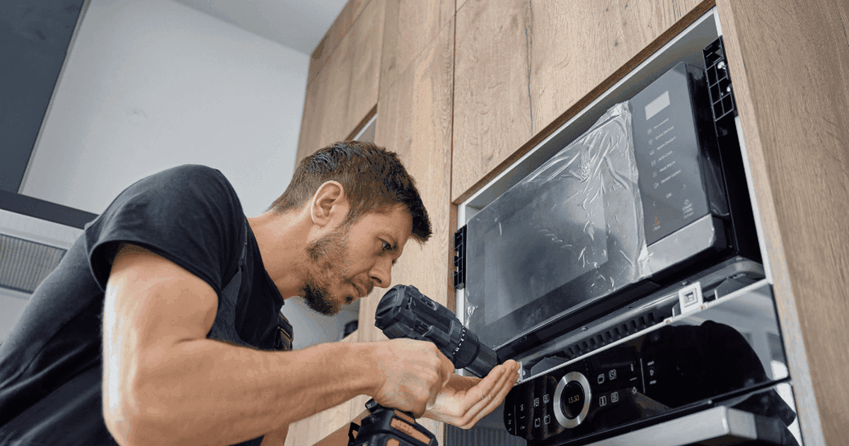 Technician using a cordless drill to fit a built-in microwave above a built-in oven in a wood-panelled kitchen