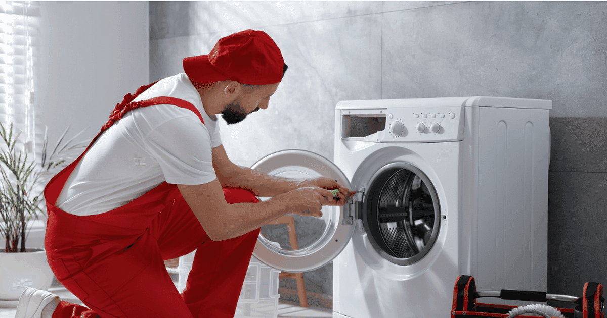 Technician in red overalls kneeling beside an open front-loading washing machine, with a toolbox and the detergent drawer on the floor