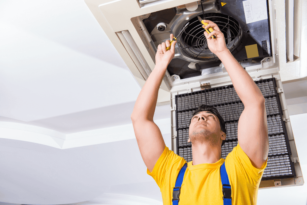 Technician in a yellow shirt inspecting the fan and filter of an opened ceiling cassette air conditioner