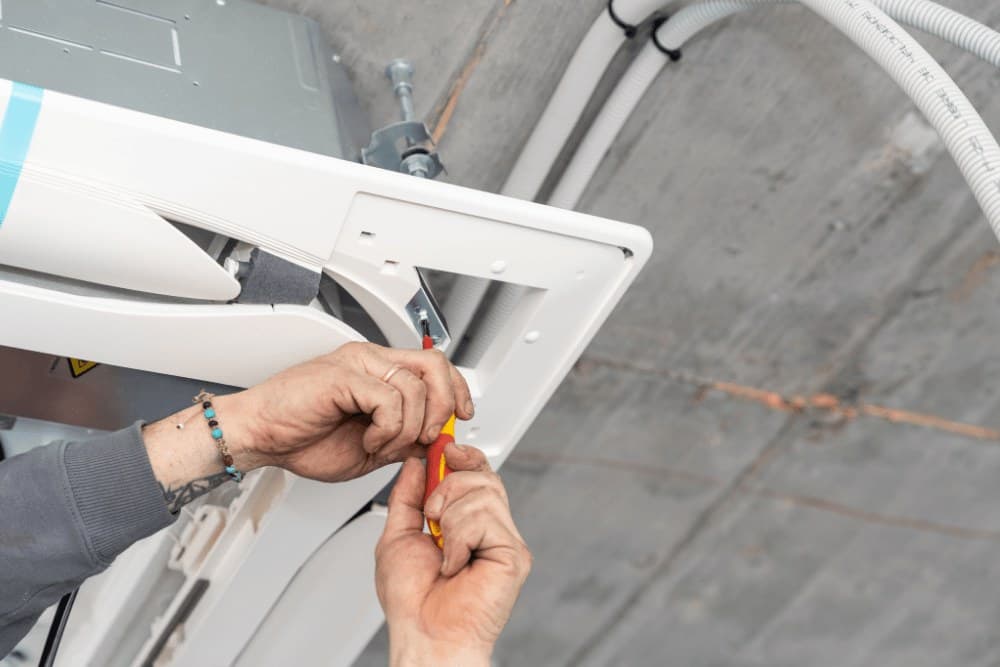 Hands using a screwdriver to fix the front panel of a ceiling cassette indoor unit during a Mitsubishi Electric AC service