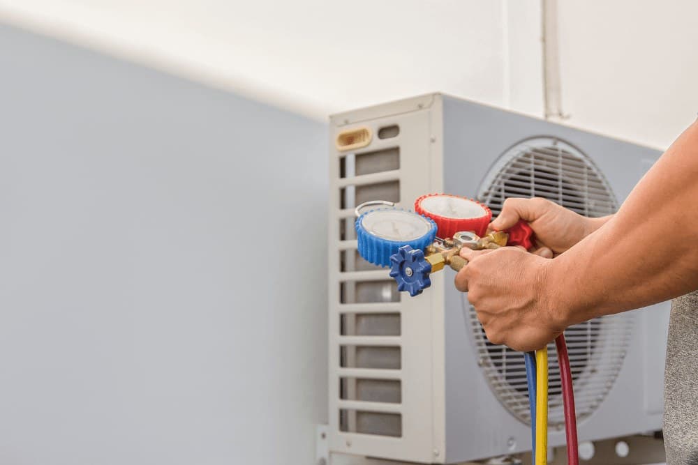 Hands holding a red and blue manifold gauge set connected to an outdoor split air conditioner unit