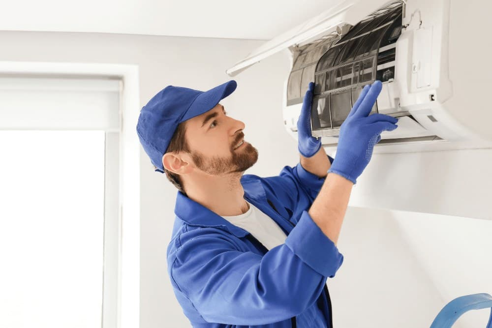 Technician in a blue cap and gloves lifting the front panel of a wall-mounted split AC indoor unit, the typical Gree split-system service