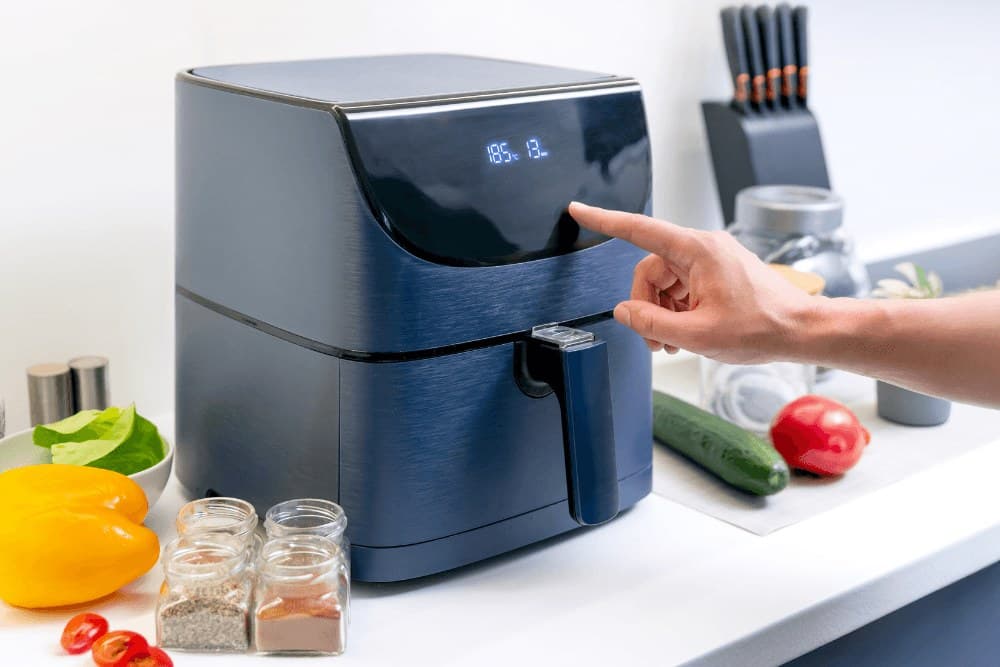 Close-up of a finger pressing the digital touch display of a navy-blue air fryer on a kitchen counter, with fresh vegetables visible beside it