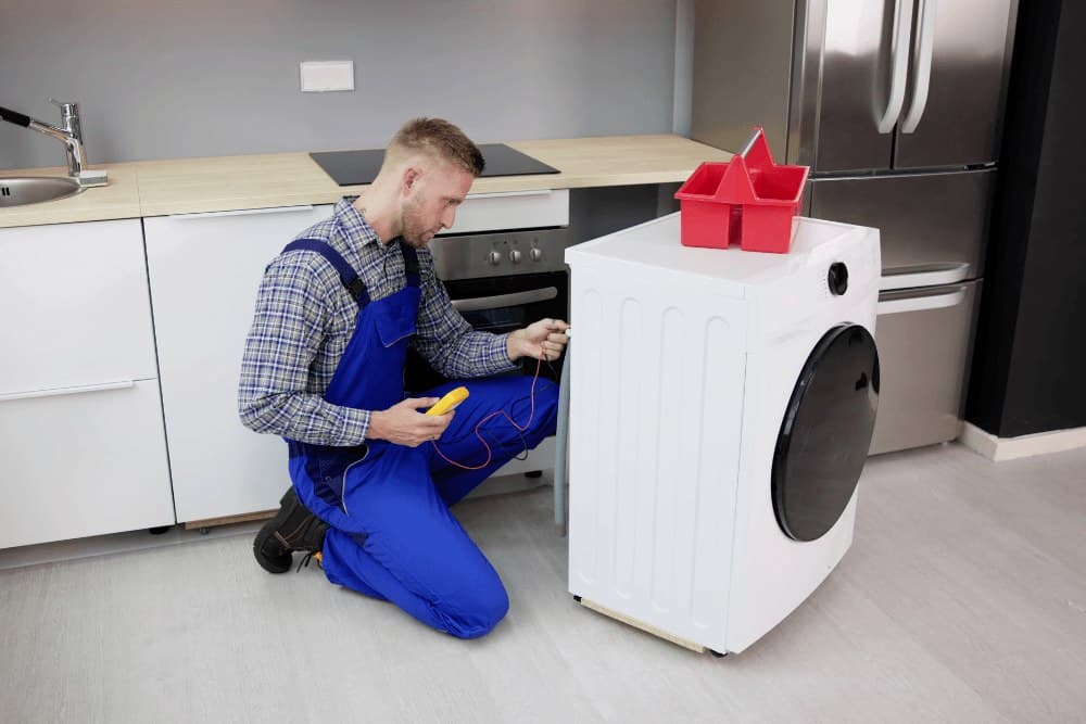 Technician in plaid shirt and blue overalls kneeling in a kitchen with a multimeter in hand, testing a built-in oven beside a washing machine
