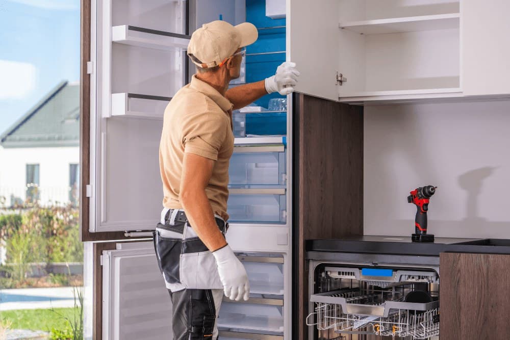 Technician in a cap and white gloves working at a built-in kitchen with a fridge column and an integrated dishwasher with its rack pulled out