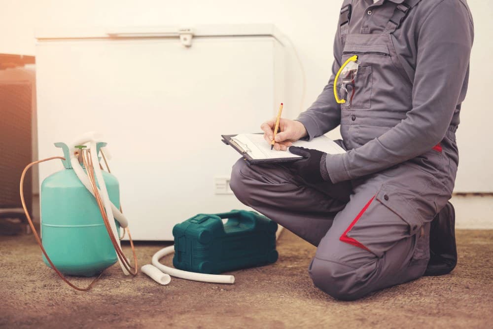 Technician in gray overalls kneeling beside a white chest freezer with a refrigerant cylinder, hoses and a teal toolbox on the floor, filling in a service checklist during a repair in Abu Dhabi