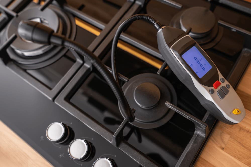 One Click Technicians digital gas leak detector resting on a black gas hob, its probe positioned beside the burner and control knobs, with the display lit up reading the area for a leak