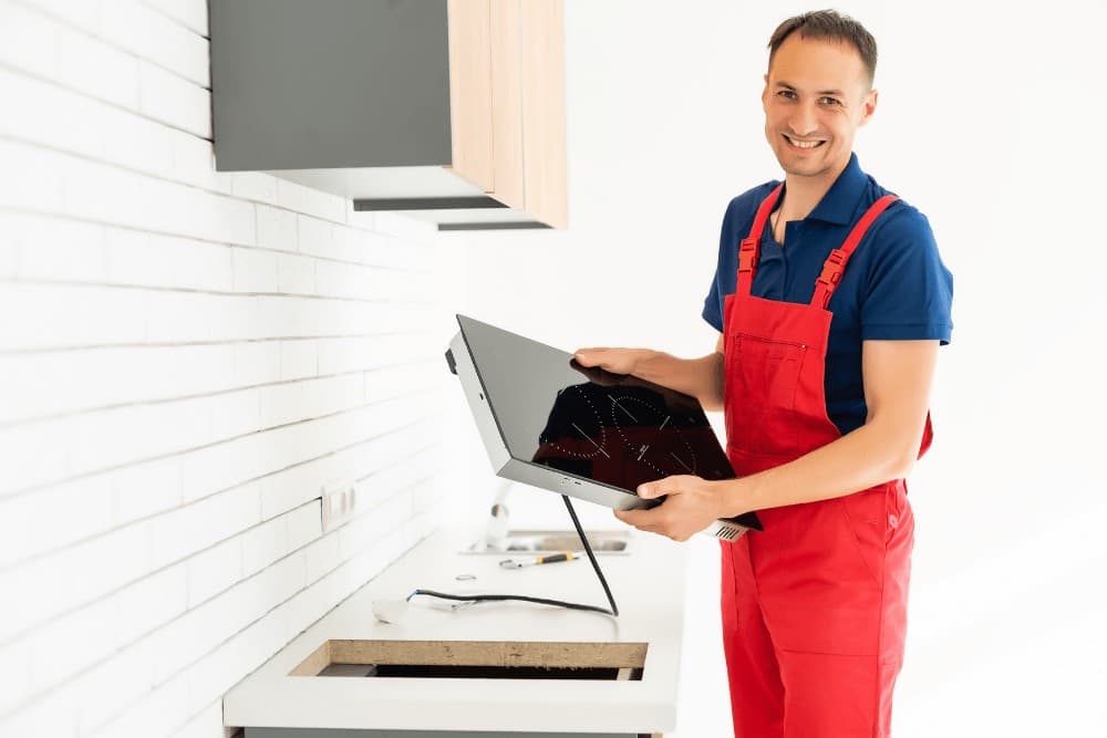 Smiling technician in red overalls holding a new induction hob at an angle before fitting it into a kitchen worktop cut-out