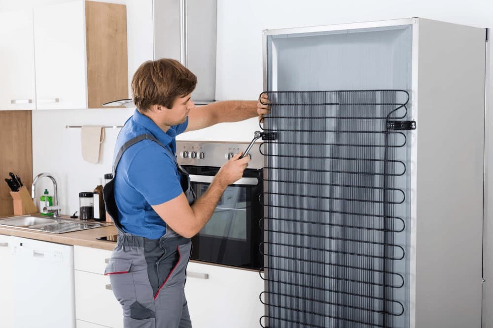 Technician in a blue polo shirt standing beside a tall built-in refrigerator column, fitting the rear condenser coil panel during a repair