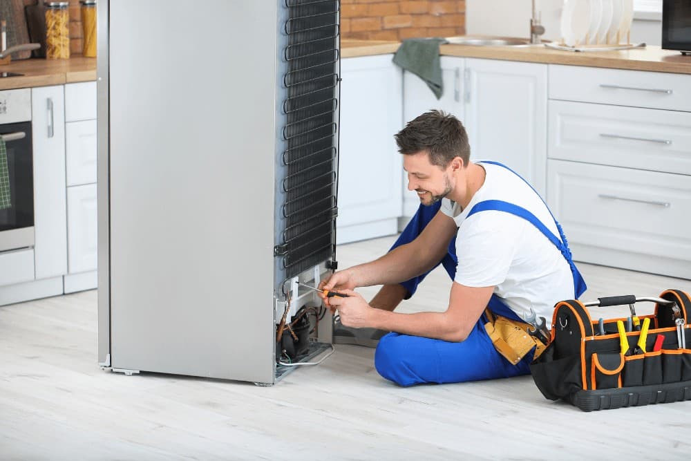 Technician in a blue polo shirt kneeling beside a refrigerator pulled away from the wall, working on its rear panel with a toolbox bag open on the floor