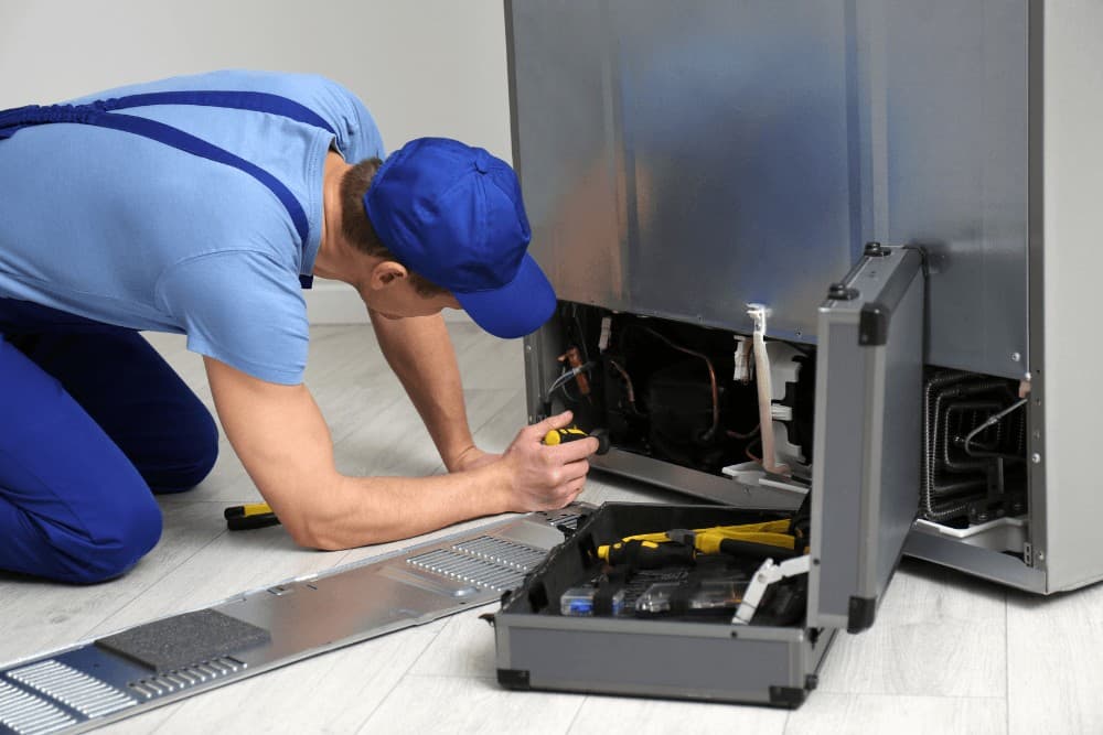 Technician in a blue cap kneeling at the base of a refrigerator with the rear panel open, working on the compressor area with a toolbox on the floor