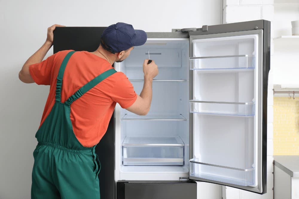 Technician in orange overalls and a blue cap reaching into an open, empty refrigerator to adjust an interior shelf during a repair