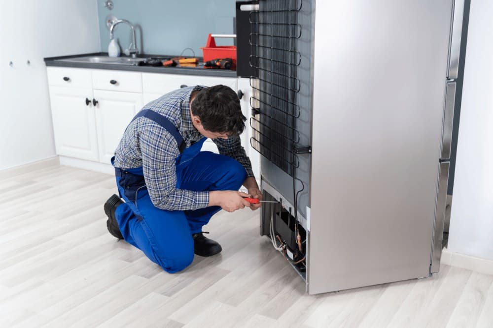 Technician in blue overalls kneeling at the back of a refrigerator, working on the condenser coil and compressor area