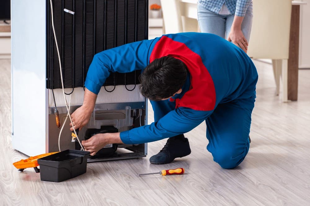 Technician in blue and red overalls kneeling at the back of a black refrigerator, working at its base with a toolbox and screwdriver on the floor