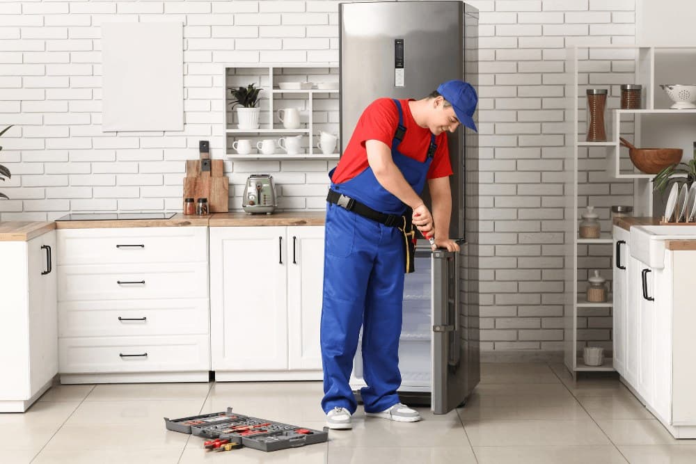 Technician in a red shirt and blue overalls with a tool belt working on the door of a tall stainless refrigerator in a white-tiled kitchen