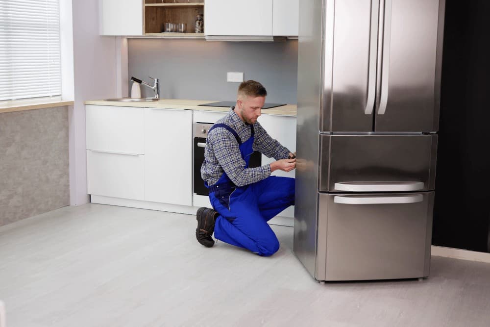 Technician in a plaid shirt and blue overalls kneeling beside a tall stainless steel refrigerator, working on its lower front panel