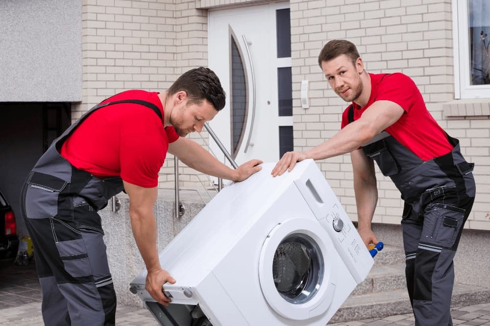 Two technicians in red shirts and gray work trousers carrying an old washing machine together out of a property for removal and disposal
