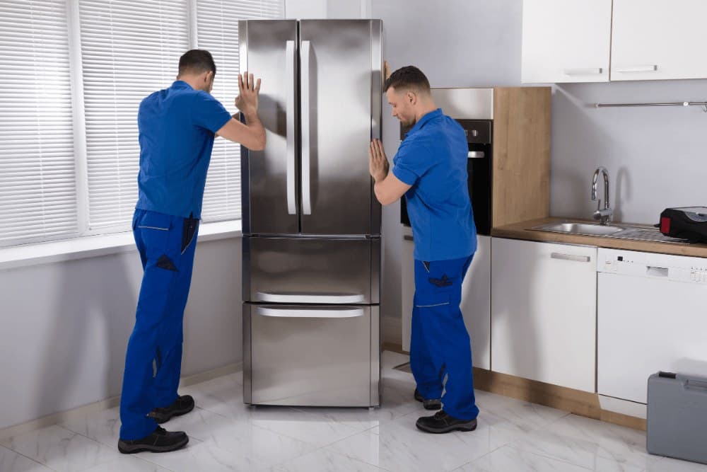 Two technicians in blue overalls moving a stainless steel French-door refrigerator out of a kitchen