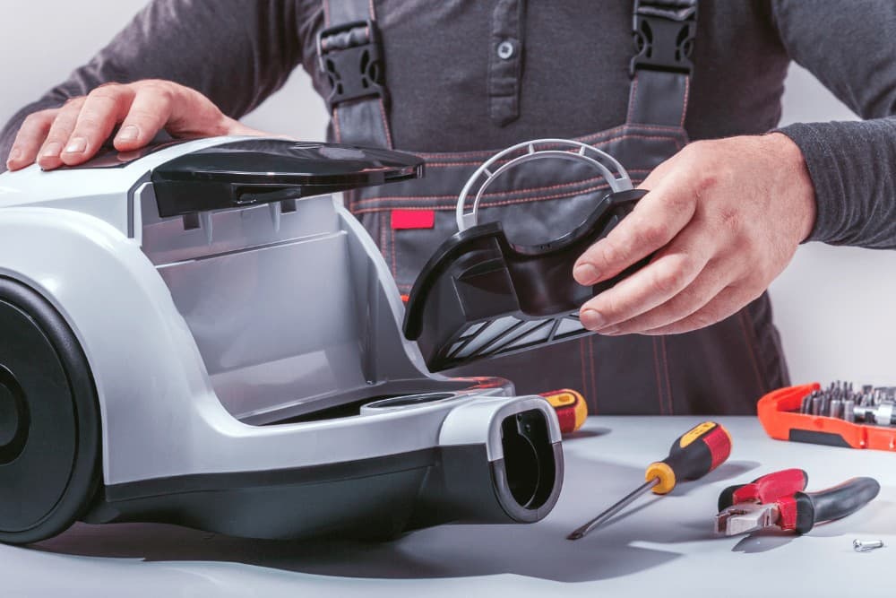 Hands reassembling a gray vacuum cleaner's dust canister and motor housing on a workbench, with a toolbox and spare parts laid out beside the unit