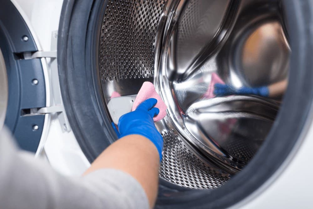 Gloved hand wiping the inside of a front-loading washing machine drum with a pink cloth during a routine clean