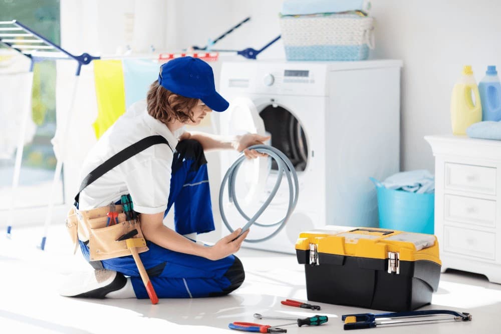 Technician in blue cap kneeling beside a newly positioned washing machine, connecting the inlet hose as part of a new installation in a laundry room