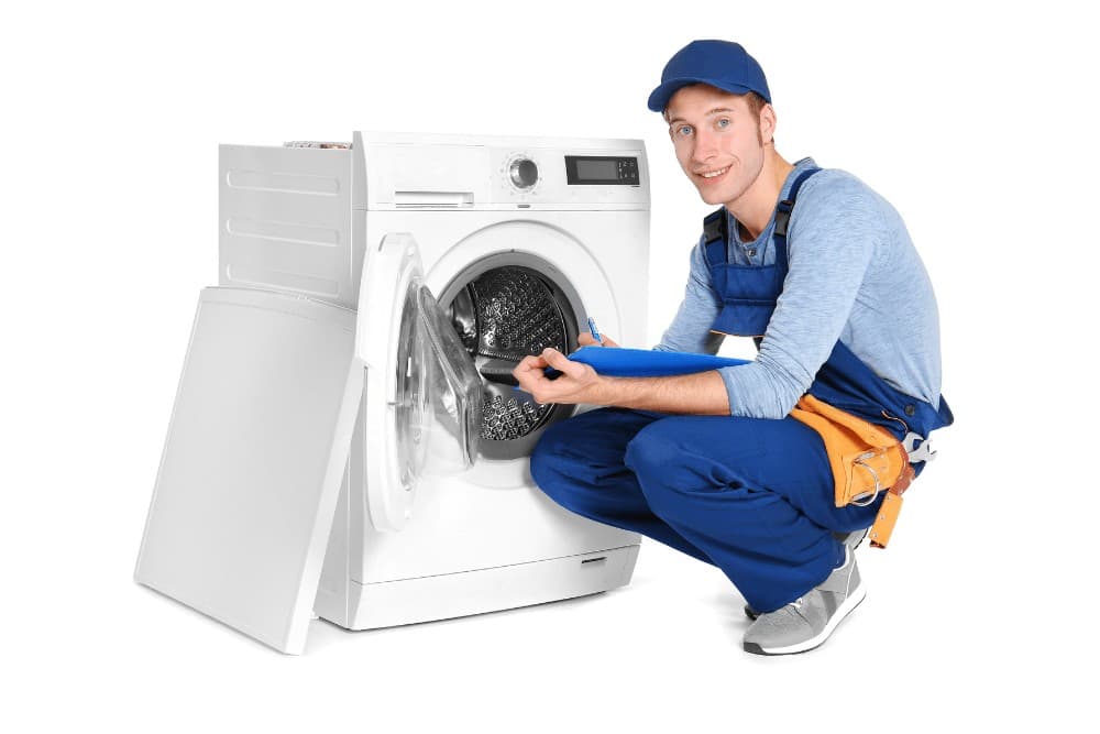 Smiling technician in a blue cap and overalls crouching beside an open front-loading washing machine, holding a blue clipboard