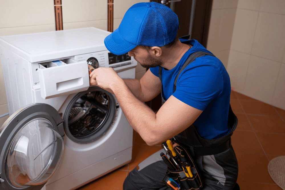 Technician in a blue cap working on a front-loading washing machine with the detergent drawer pulled out