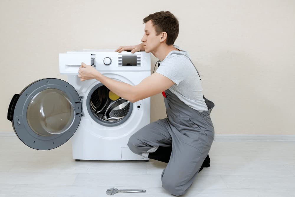 Technician in gray overalls kneeling in front of an open front-loading washing machine, inspecting the drum during a repair