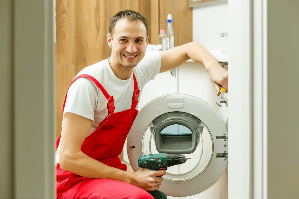 Smiling technician in red overalls kneeling beside a front-loading washing machine, holding a cordless drill