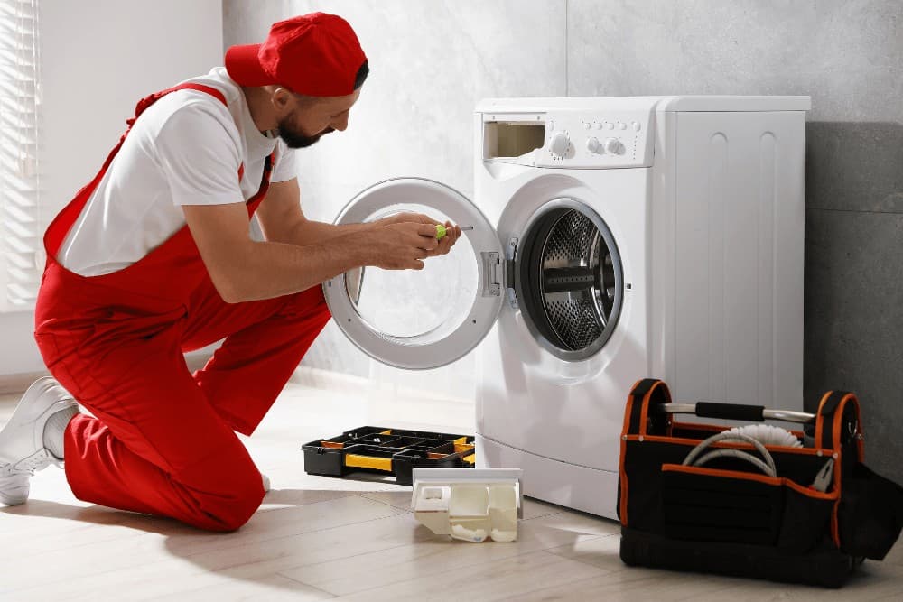 Technician in red overalls kneeling beside an open front-loading washing machine, with a toolbox on the floor during a repair