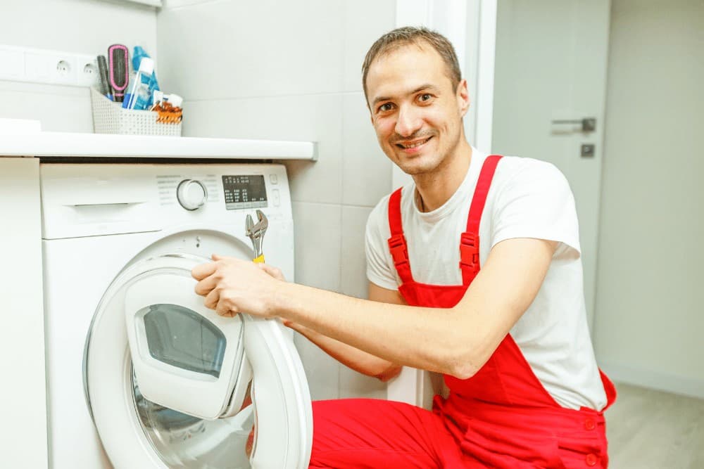 Smiling technician in red overalls holding the door of a washing machine, with a wrench in his other hand