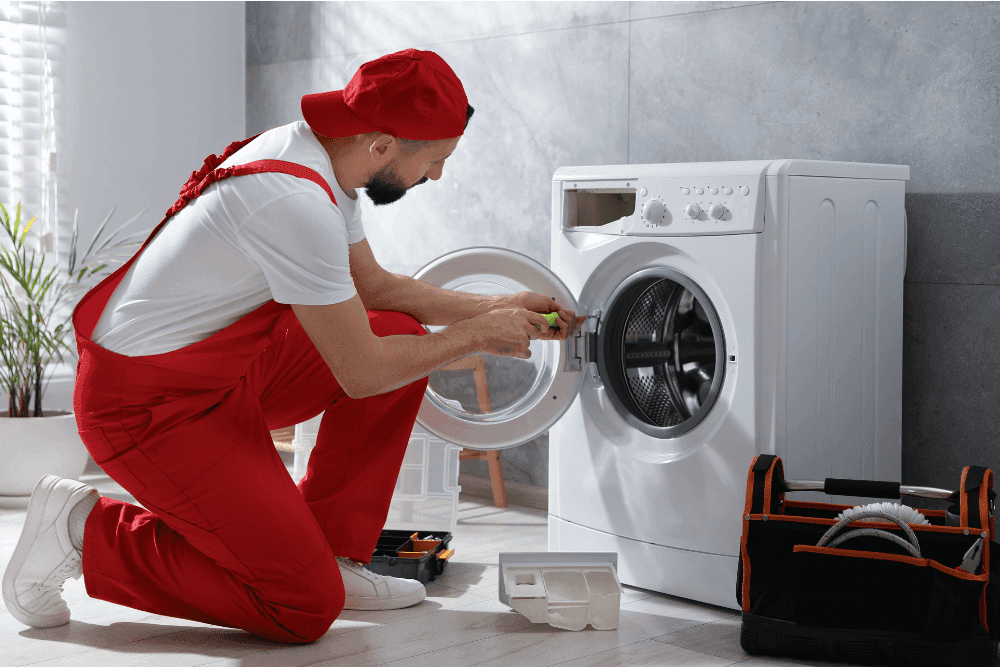 Technician in red overalls kneeling beside an open front-loading washing machine, with a toolbox and the detergent drawer pulled out