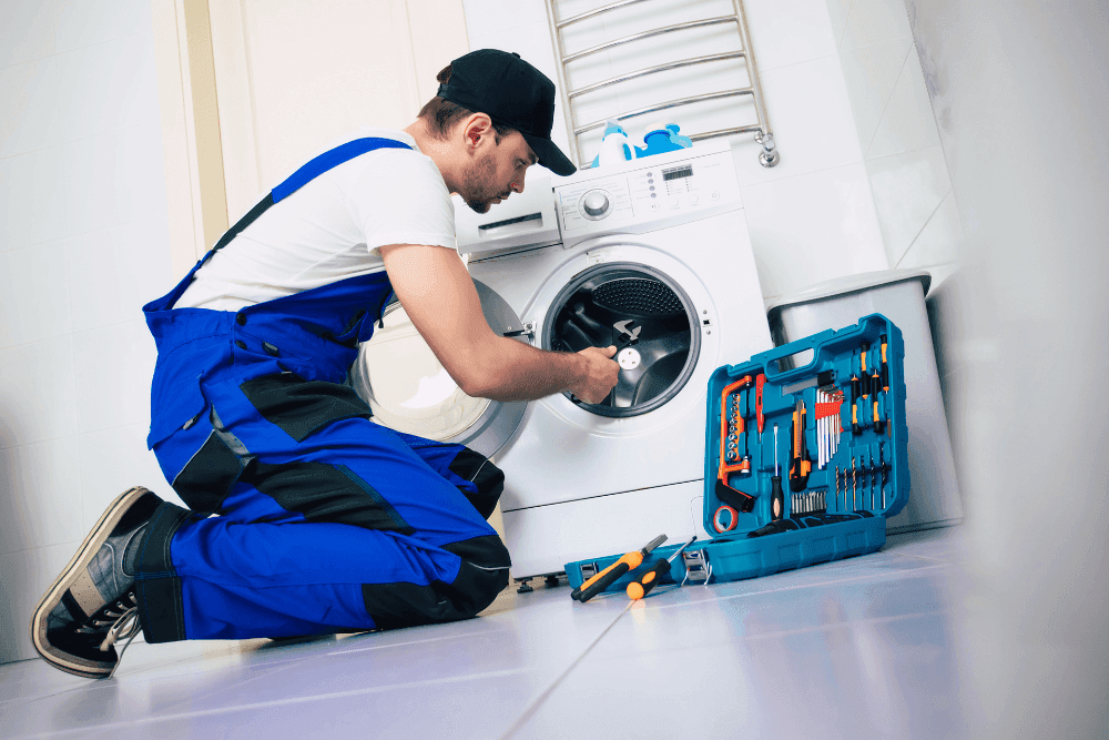 Technician in blue overalls kneeling beside an open washing machine with a teal toolbox on the floor