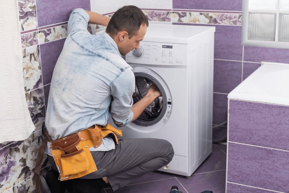 Technician in a denim shirt and tool belt kneeling in front of a washing machine in a purple-tiled bathroom, reaching into the drum