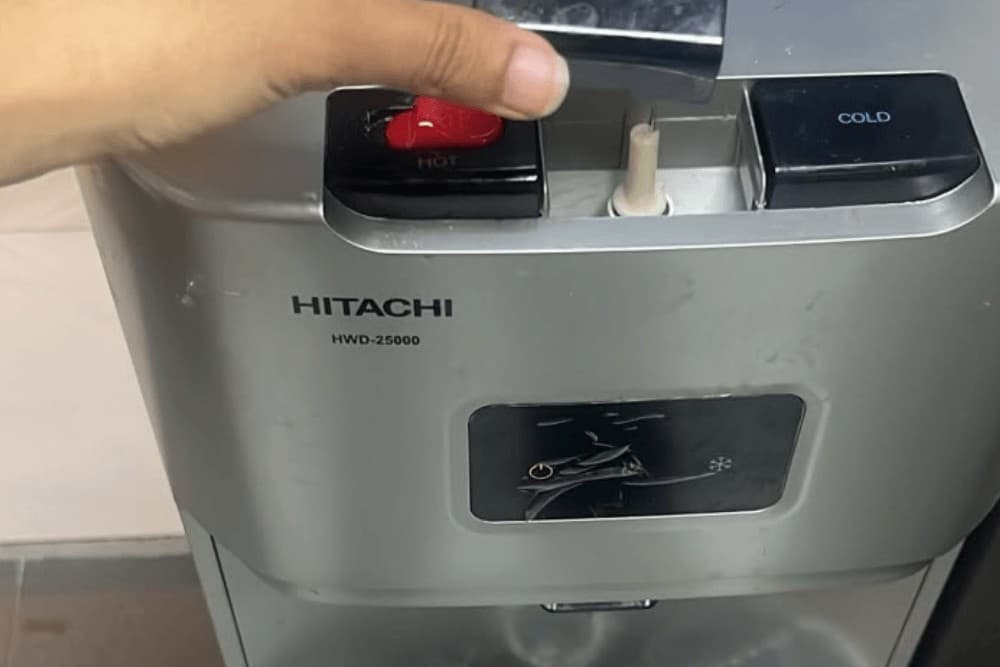 Close-up of a hand pressing the cold-water button on a water dispenser's stainless steel control panel, with the hot and cold indicator lights visible