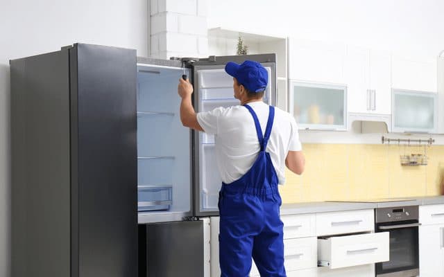 Technician in blue overalls checking the inside of an open French-door refrigerator in a modern kitchen