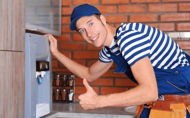 Technician in a striped shirt and cap giving a thumbs-up beside a wall-mounted water dispenser with its cold and hot taps visible, confirming the repair is complete in a kitchen